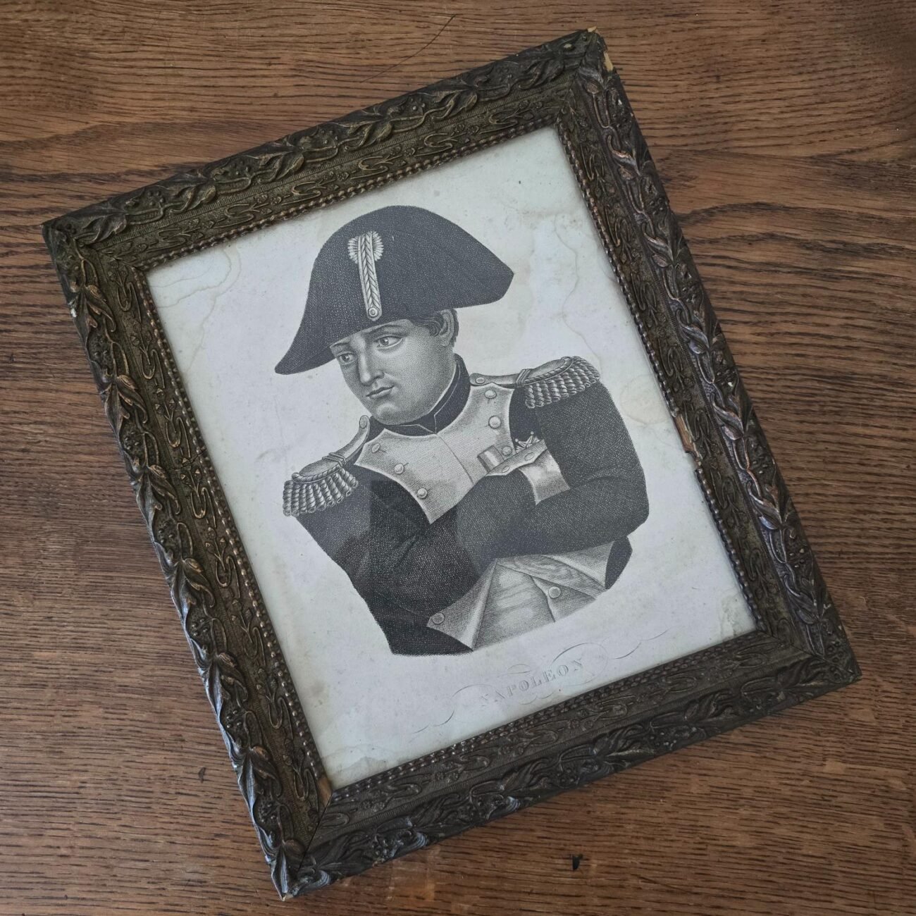 Cadre ancien sous verre représentant Napoléon Bonaparte posé sur une table en bois, en légère contre-plongée.