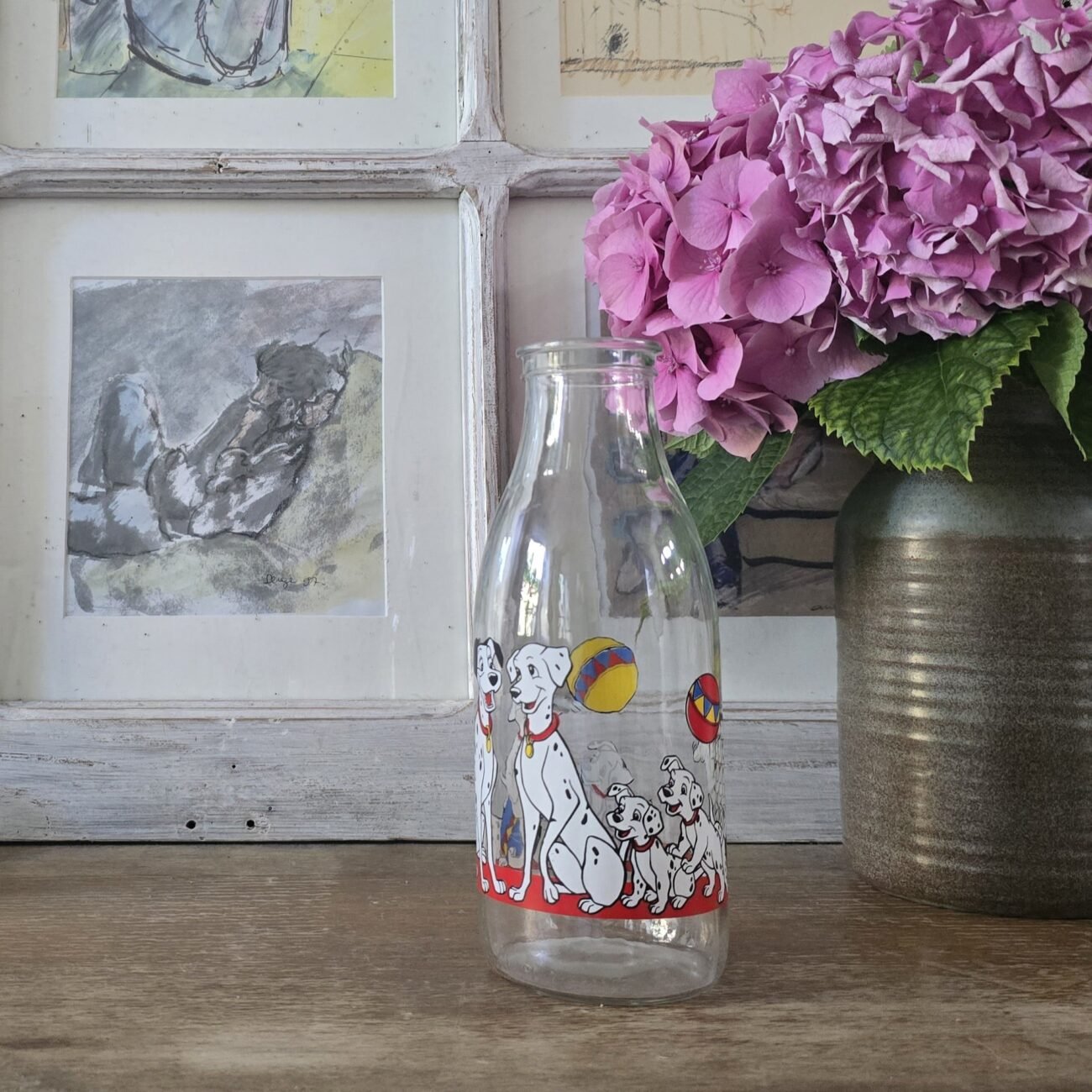 Carafe en verre décorée des 101 Dalmatiens posée sur une table en bois, à côté d’un vase fleuri et de cadres accrochés au mur.